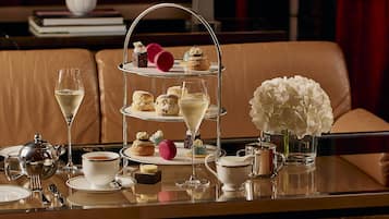 A three-tiered dessert stand with various pastries and a glass of champagne on a table in a room with a leather couch and a vase of white flowers.
