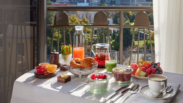 A breakfast table with various food items and drinks, a city view through a window, and a cityscape in the background. - Park Hyatt Melbourne (East Melbourne)
