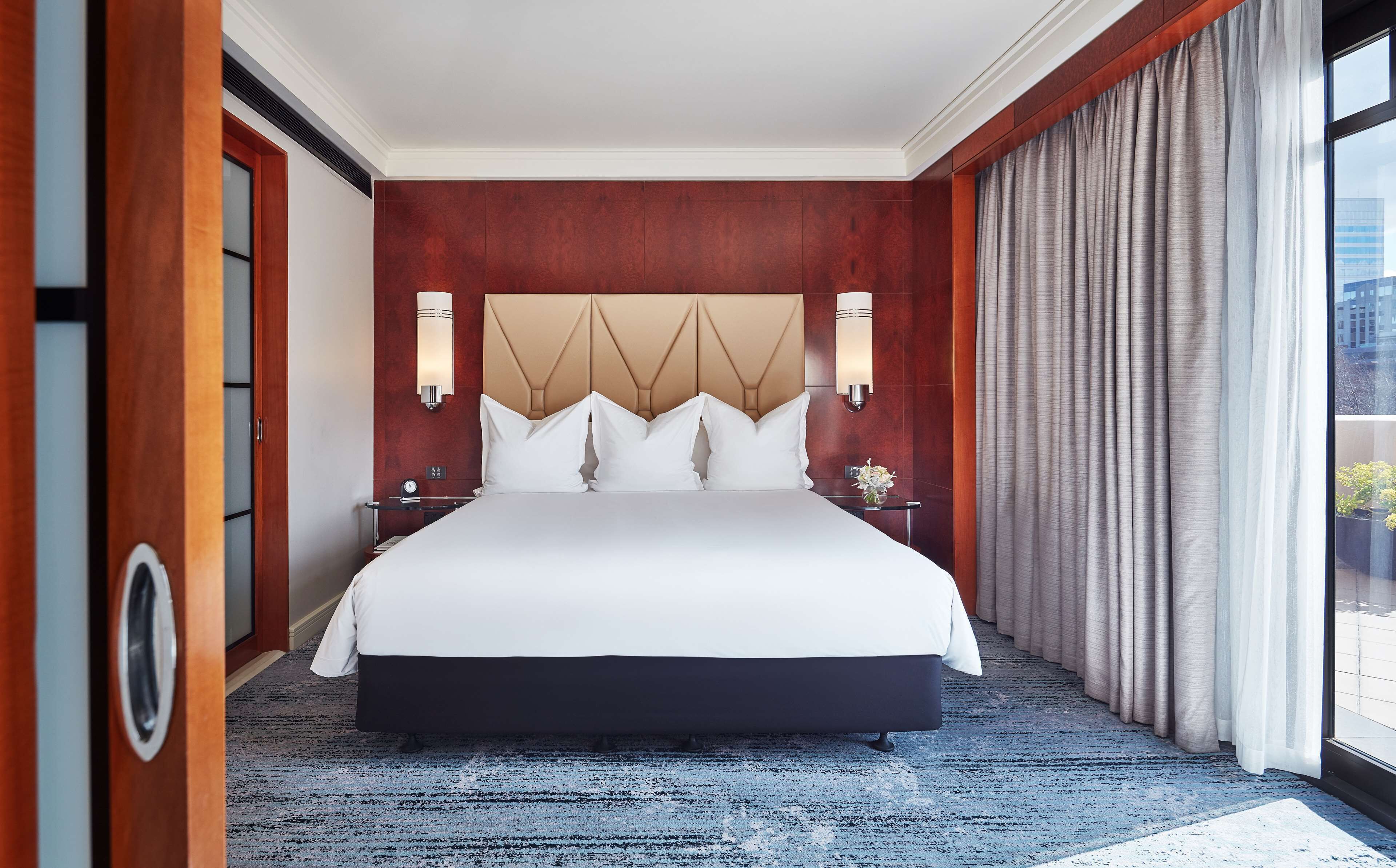 A hotel room with a large bed, wooden paneling, a modern headboard, two bedside tables with lamps, and a large window with curtains.