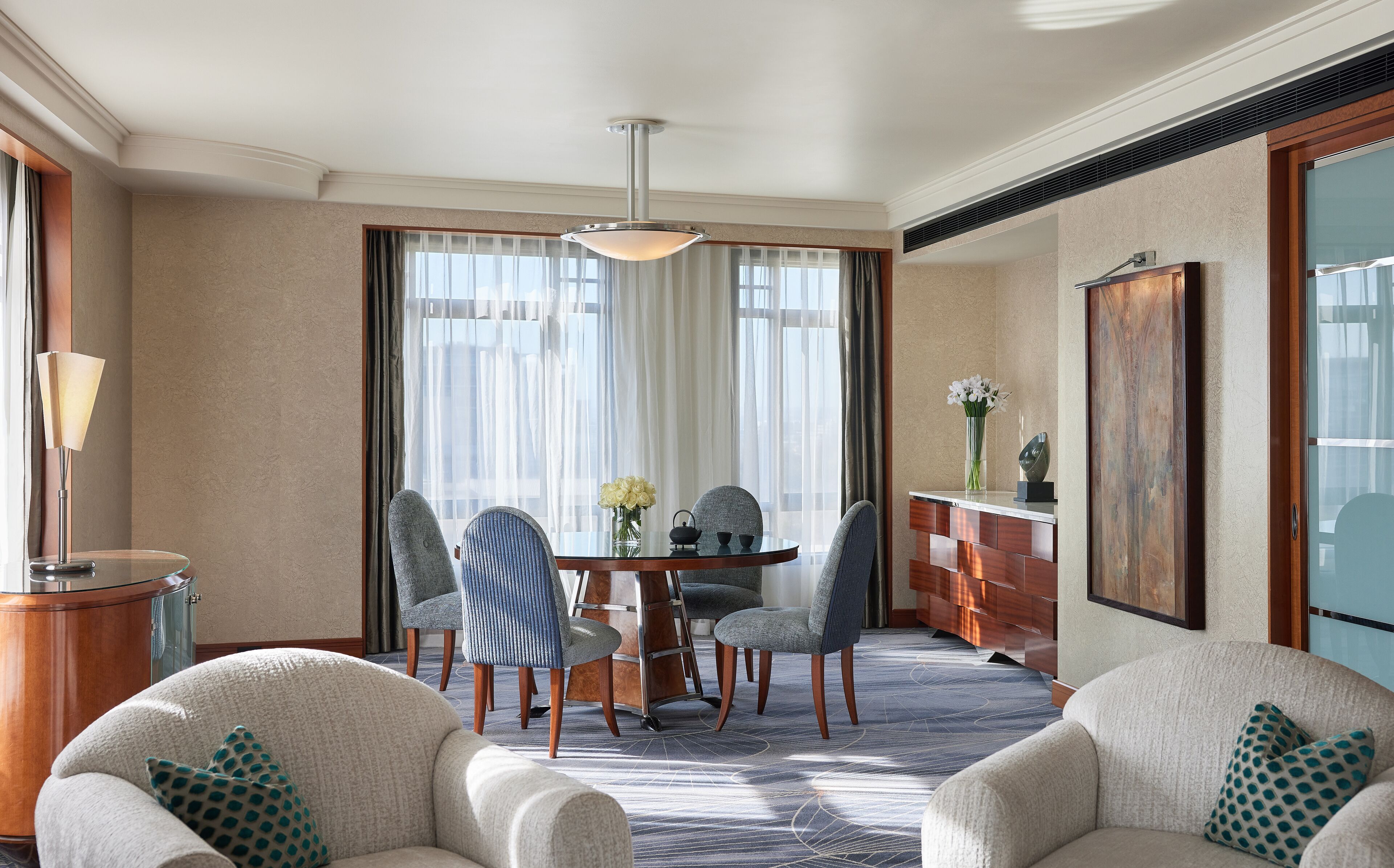 A hotel room with a round dining table, four chairs, a chandelier, a large painting, and a view of the outdoors through large windows.