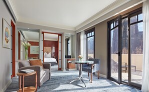 Una habitación de hotel moderna con una cama grande, una zona de estar con sofá y sillón, una mesa de comedor y un balcón con vistas a la ciudad.