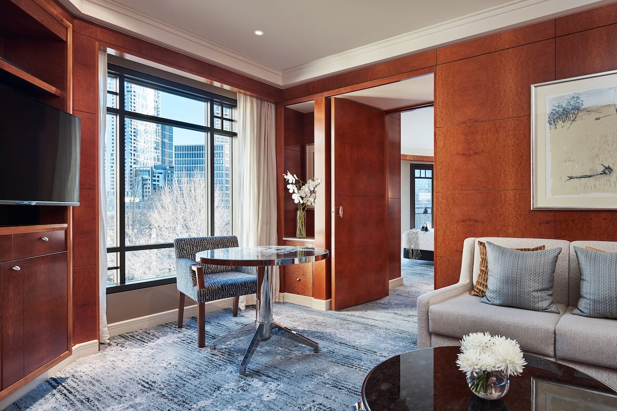 A modern hotel room with a large window, a flat-screen TV, a dining area with a table and chairs, a sofa with decorative pillows, and a framed artwork on the wall.