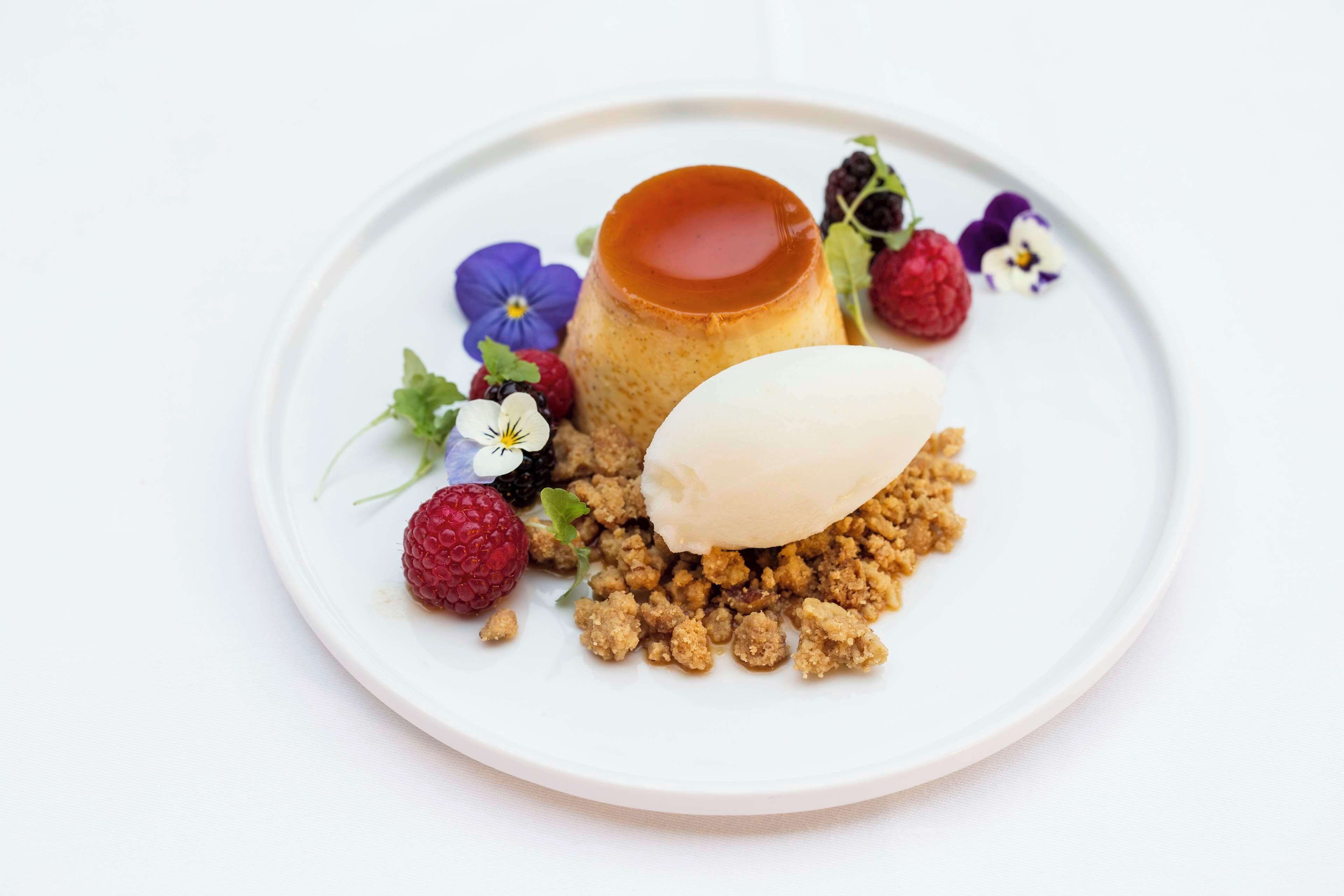 Un plato de postre con flan de caramelo, helado y cobertura desmenuzada, acompañado de frambuesas y flores.