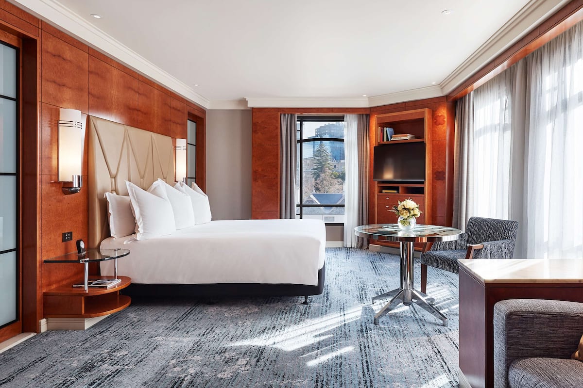 A hotel room with a large bed, a small round table with a vase, a chair, a bookshelf, and a large window with a city view.