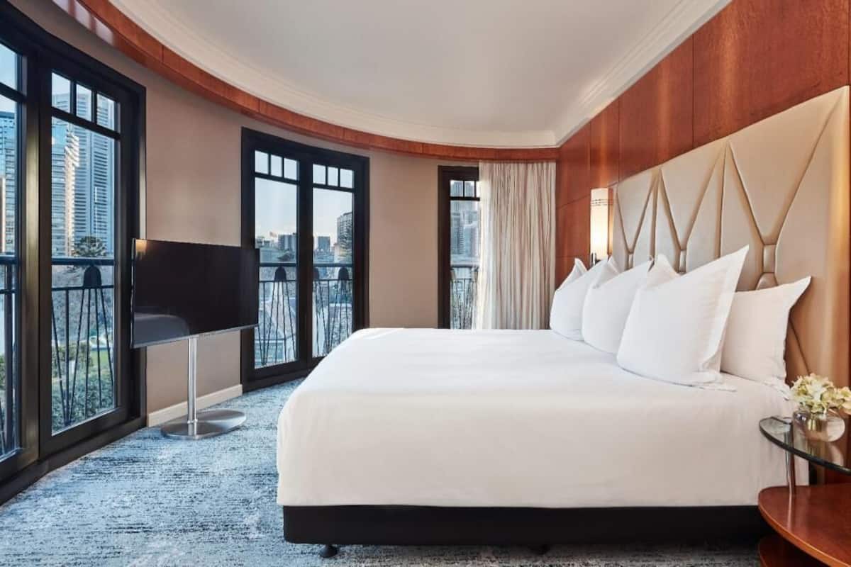Quarto de hotel com uma cama grande, televisão, mesinha e vista da cidade por grandes janelas.
