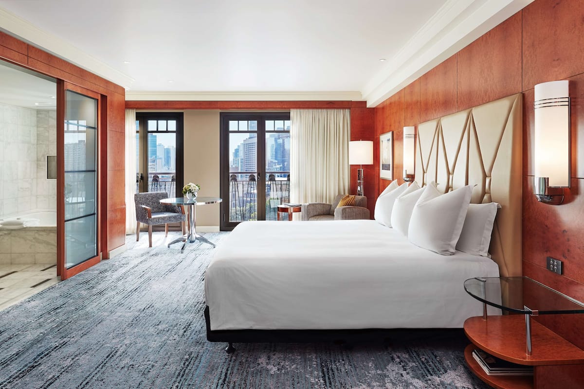 A hotel room with a large bed, a balcony with a city view, a small table with chairs, and a glass-enclosed shower area.