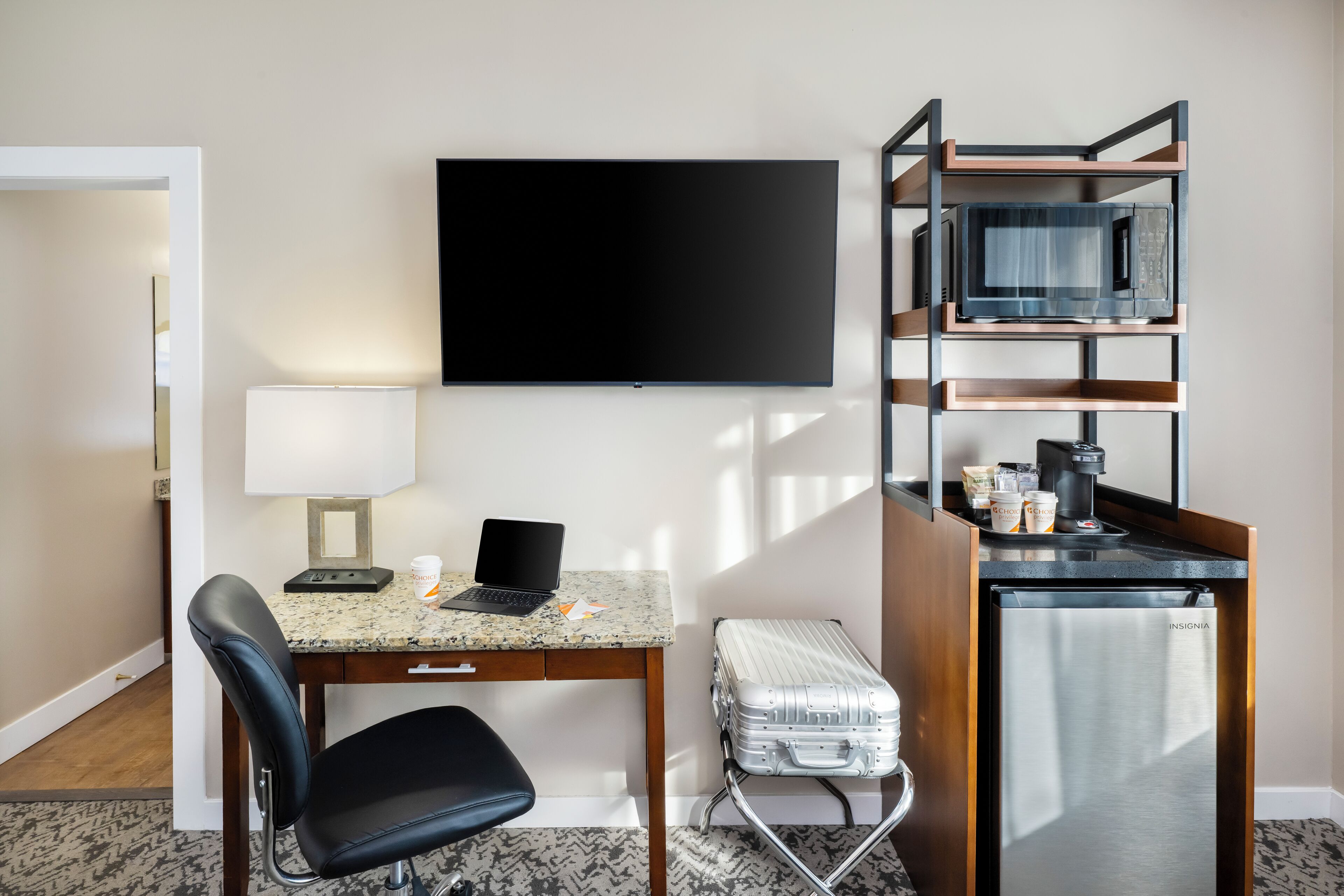 A hotel room with a flat-screen TV, a desk with a laptop, a chair, a suitcase, a mini-fridge, and a coffee machine.