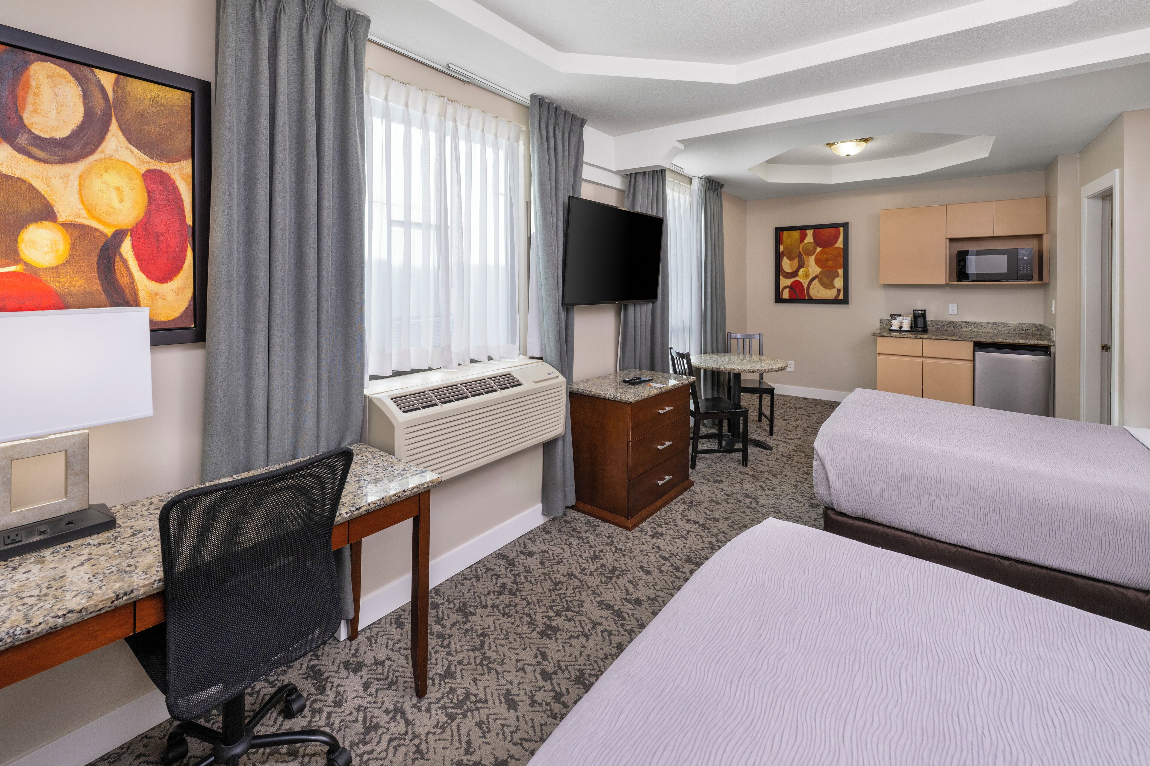 Une chambre d’hôtel avec deux lits, un bureau avec un ordinateur, une télévision, un coin cuisine et des œuvres d’art accrochées aux murs.