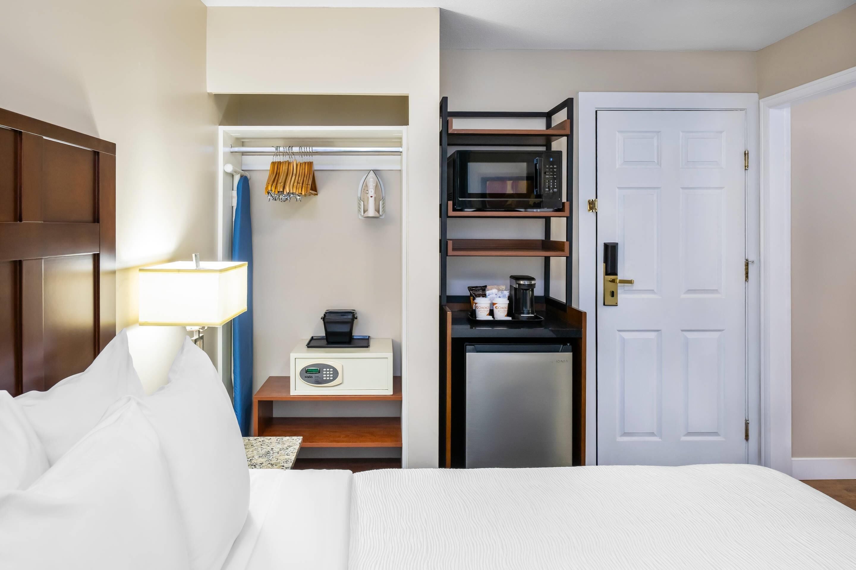 A hotel room with a bed, a desk with a microwave, a refrigerator, a closet with hanging clothes, and a lamp.