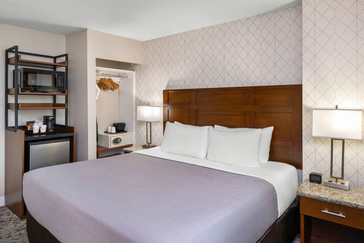 Habitación de hotel con una cama grande, un escritorio con microondas, un refrigerador pequeño, una mesita de noche con lámpara y un armario con ropa colgada.