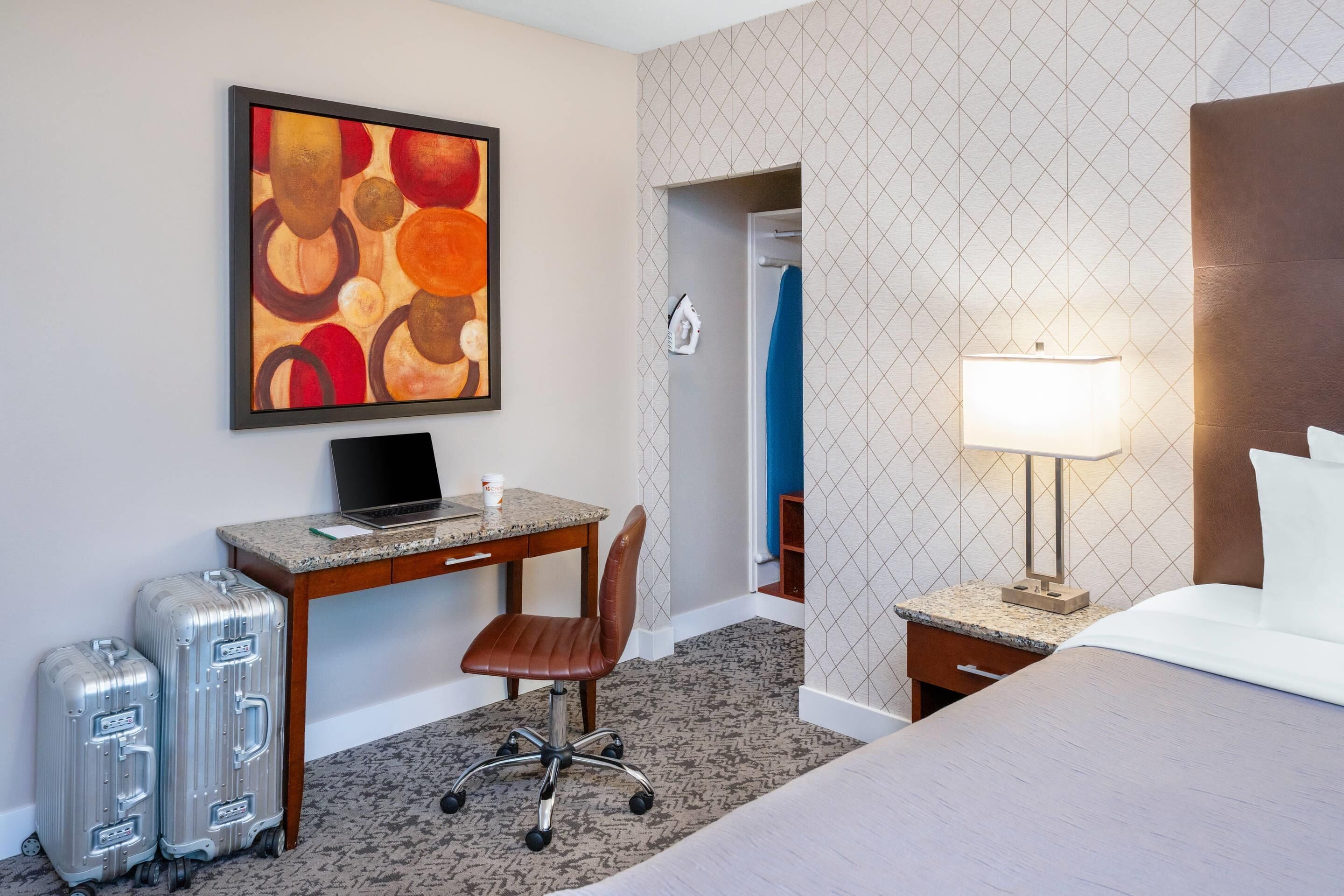 A hotel room with a bed, desk, chair, two suitcases, a lamp, and a painting on the wall.