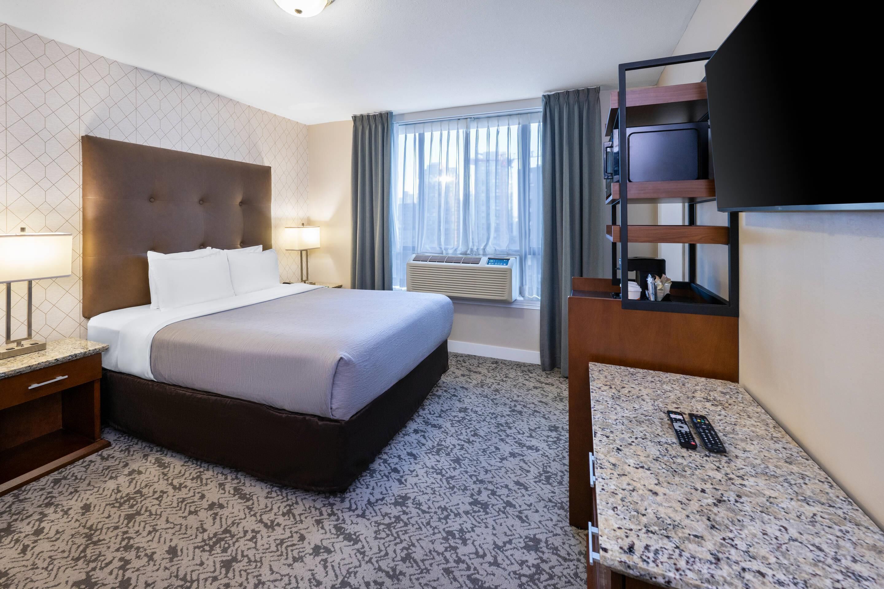 Habitación de hotel con cama, lámparas de noche, televisión, escritorio con control remoto y ventana con cortinas.