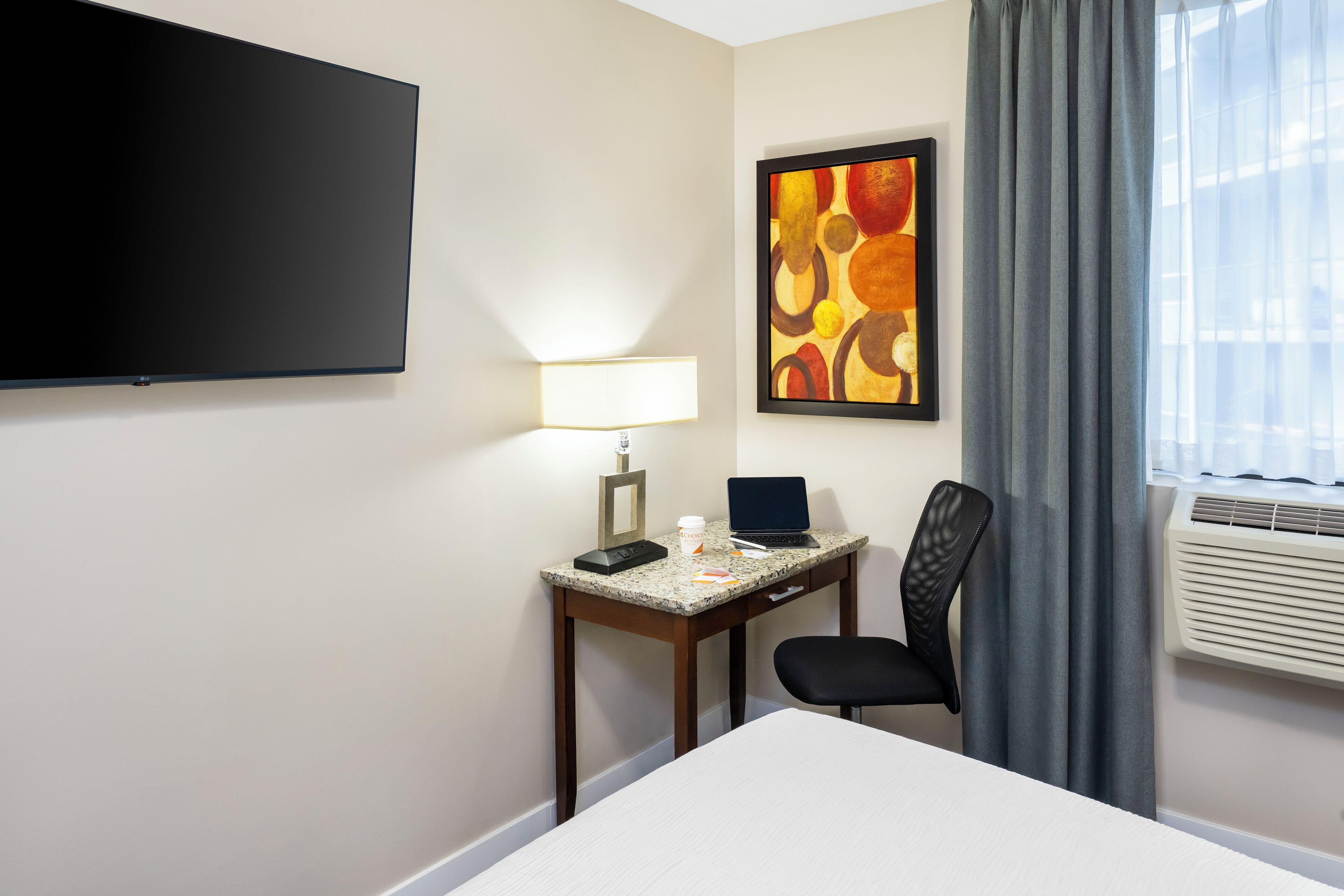 Una habitación de hotel con una cama, un escritorio con una computadora portátil, una silla, un televisor de pantalla plana, una lámpara, una obra de arte enmarcada y un equipo de aire acondicionado.