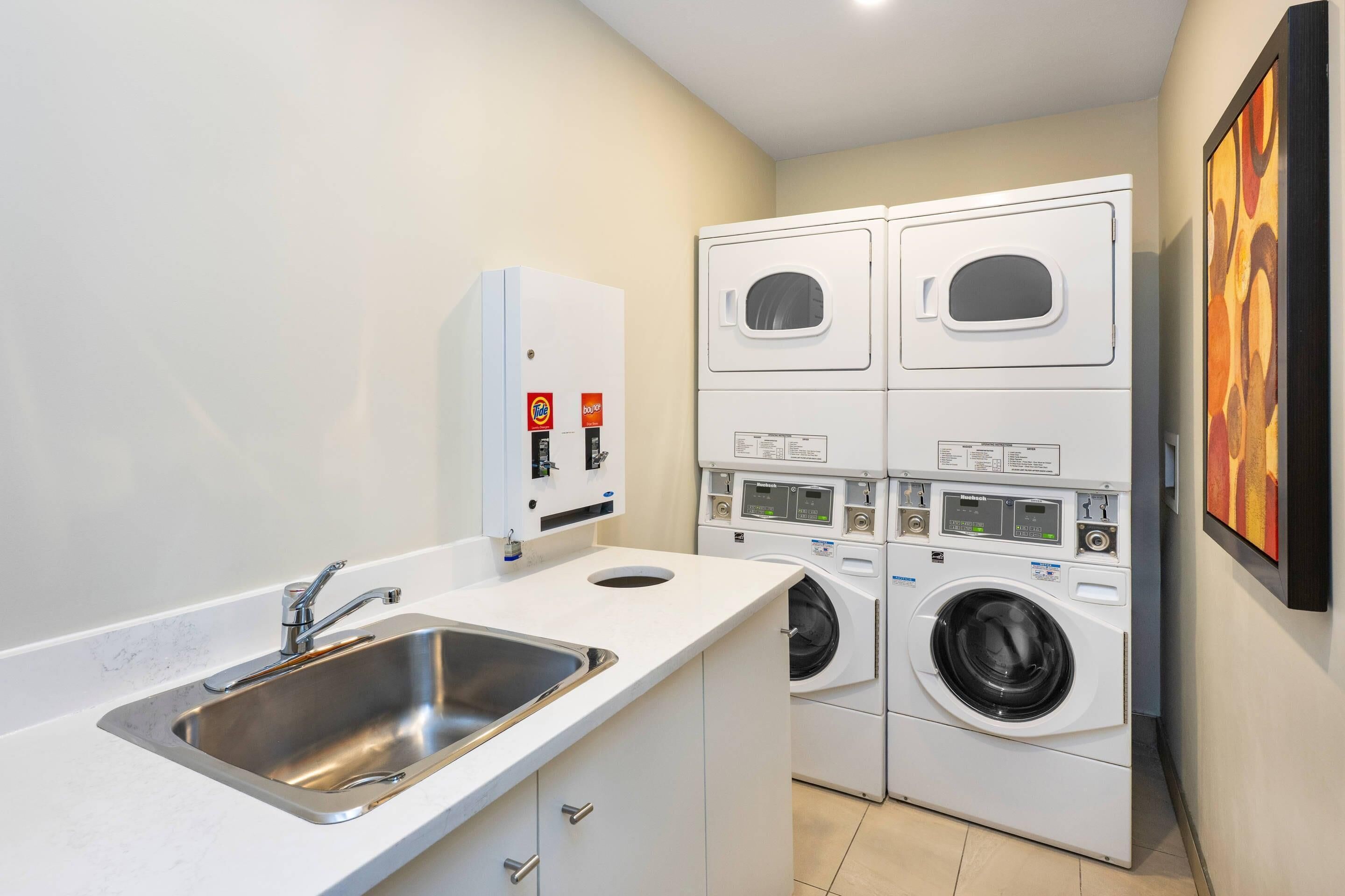 A compact laundry room with a sink, stacked washers and dryers, and a wall-mounted heater.