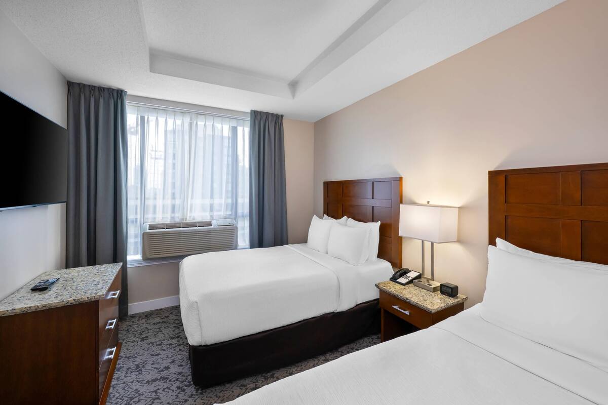 Habitación de hotel con dos camas, televisión, escritorio, lámpara y ventana con cortinas.