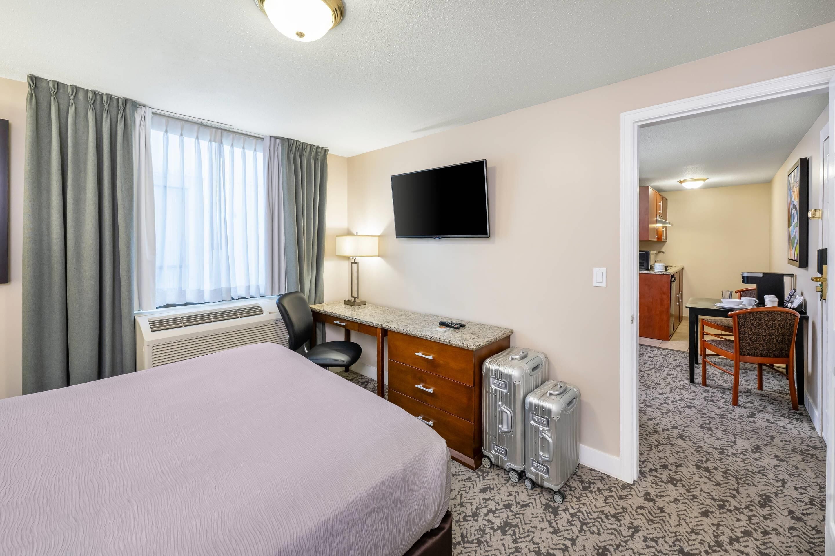 A hotel room with a bed, desk, chair, TV, and a view of the kitchen.
