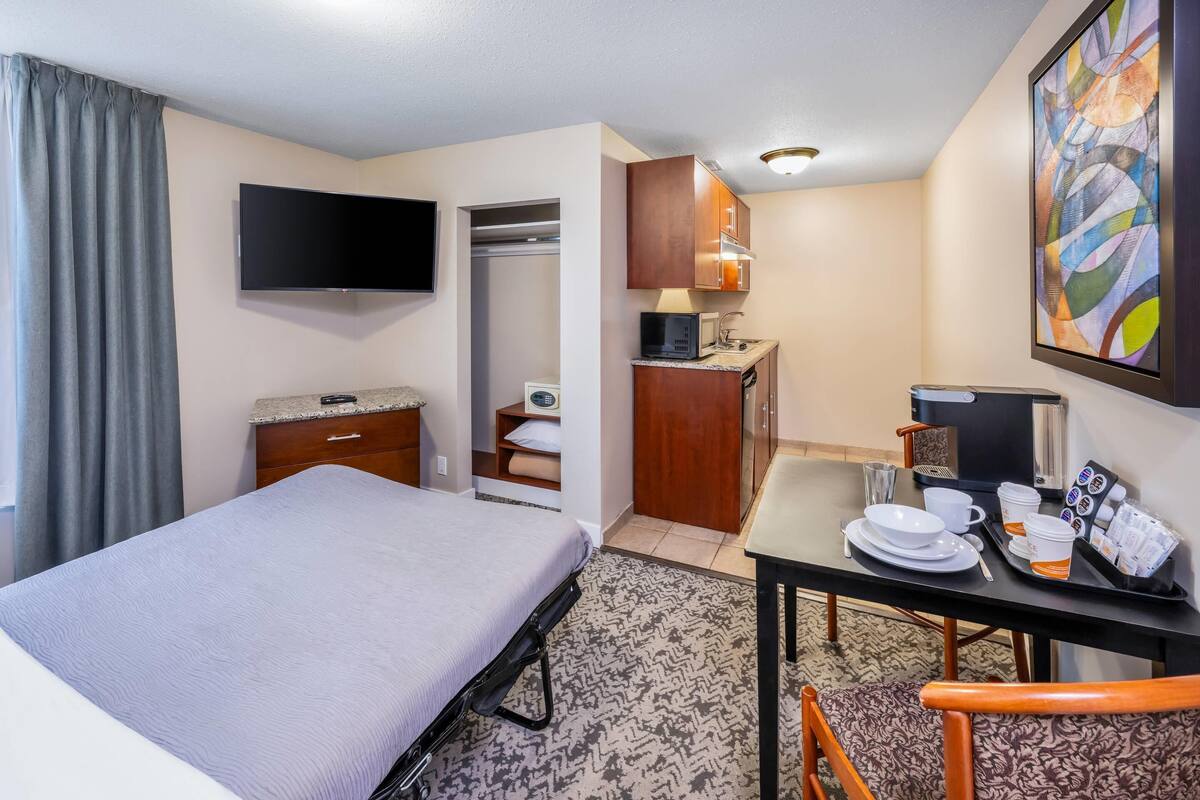 Una habitación de hotel con cama, escritorio y silla, televisión, microondas, pequeña cocina y un cuadro en la pared.
