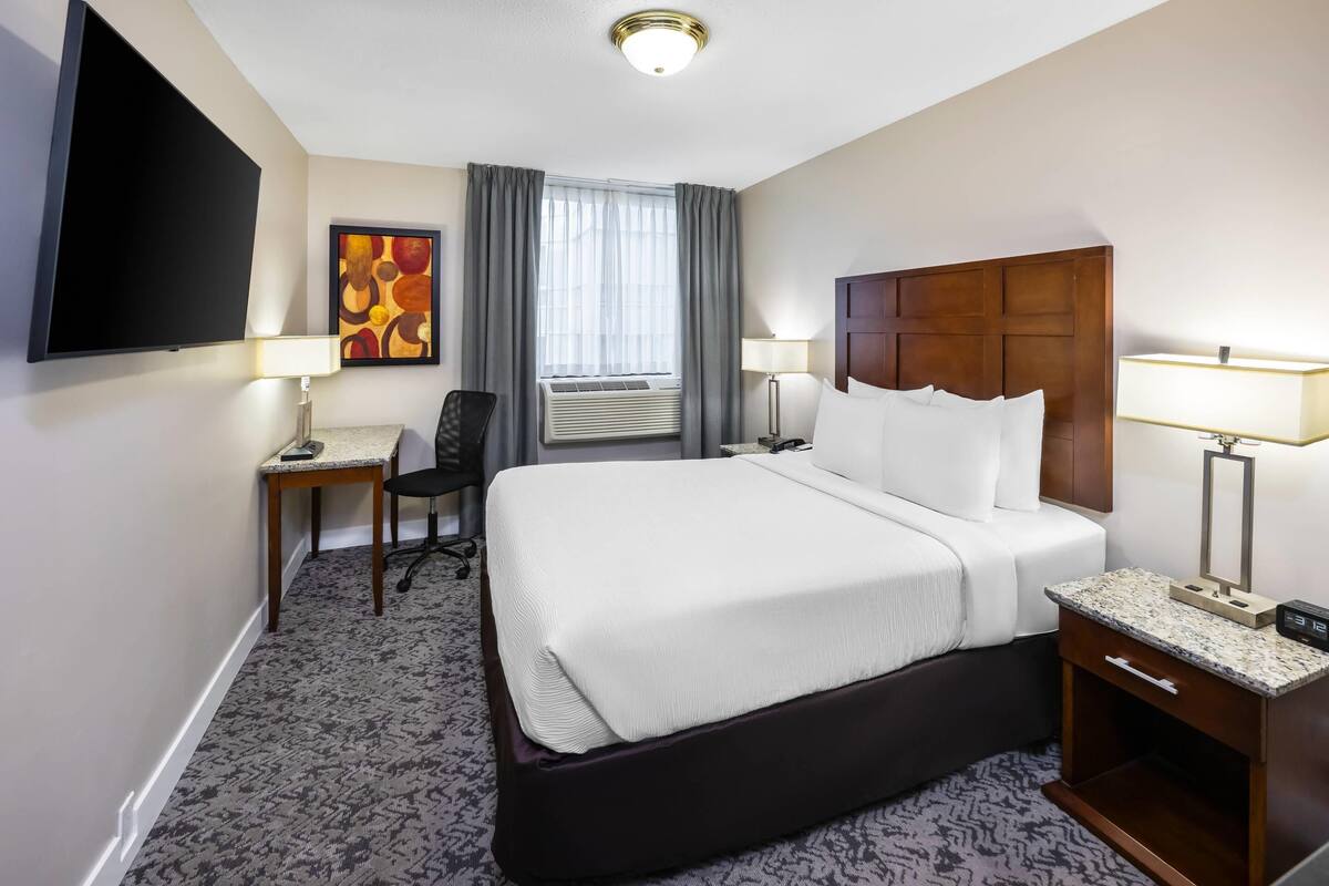 Habitación de hotel con cama, escritorio, silla, televisión, ventana con cortinas y un cuadro en la pared.