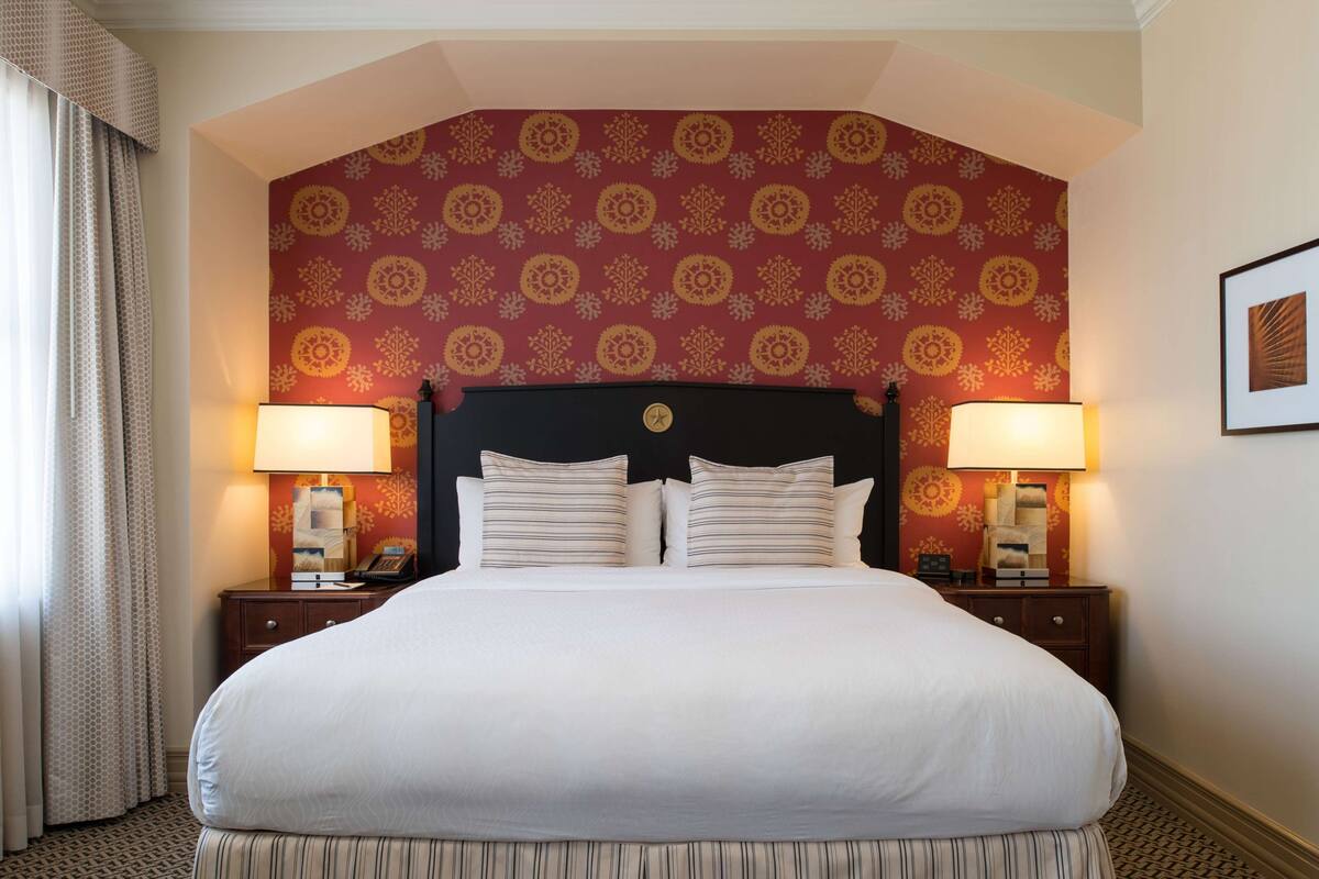 Una habitación de hotel con una cama grande, dos mesitas de noche con lámparas, y un papel tapiz estampado.