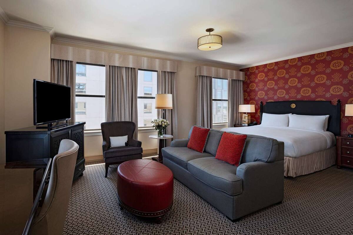 Una habitación de hotel con cama, sofá, televisor, escritorio y un reposapiés rojo.