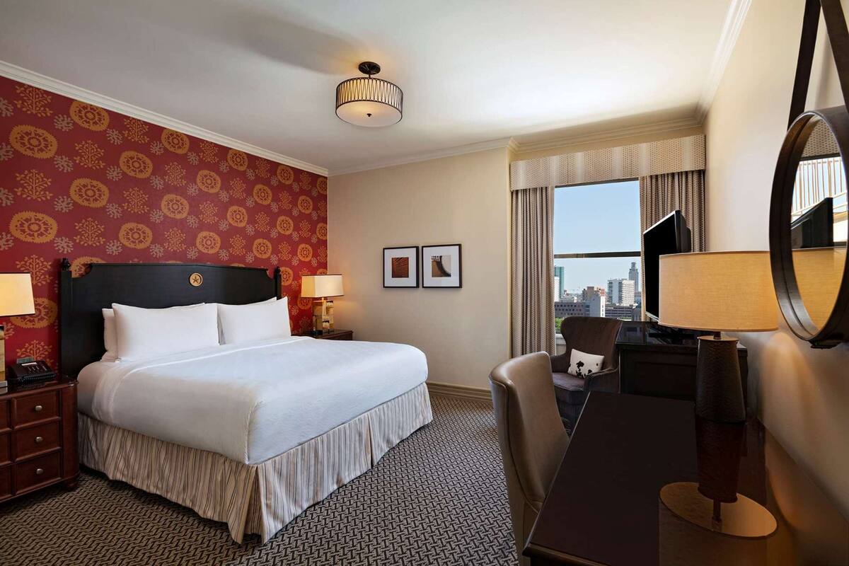 Habitación de hotel con una cama grande, un escritorio, una silla, una lámpara de escritorio, un espejo y vistas a la ciudad desde la ventana.