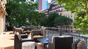 Un área de estar al aire libre con mobiliario de mimbre y una pequeña mesa, rodeada de árboles y con vistas a un paisaje urbano con edificios modernos e históricos.