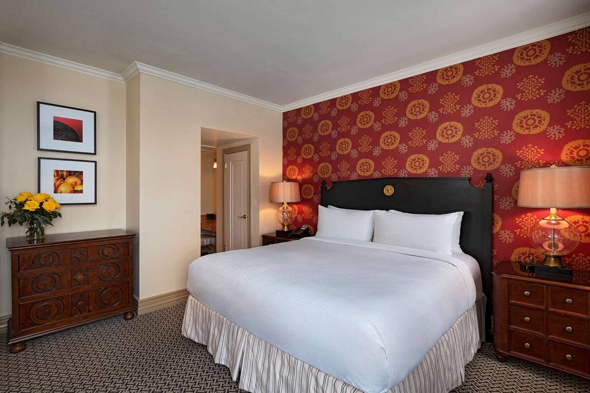Una habitación de hotel con una cama grande, dos mesitas de noche, un aparador y un papel tapiz estampado.
