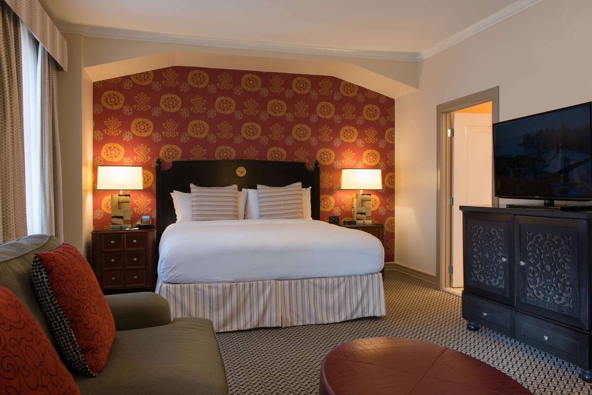 Habitación de hotel con una cama grande, dos mesitas de noche con lámparas, un televisor sobre un mueble y un papel tapiz estampado.