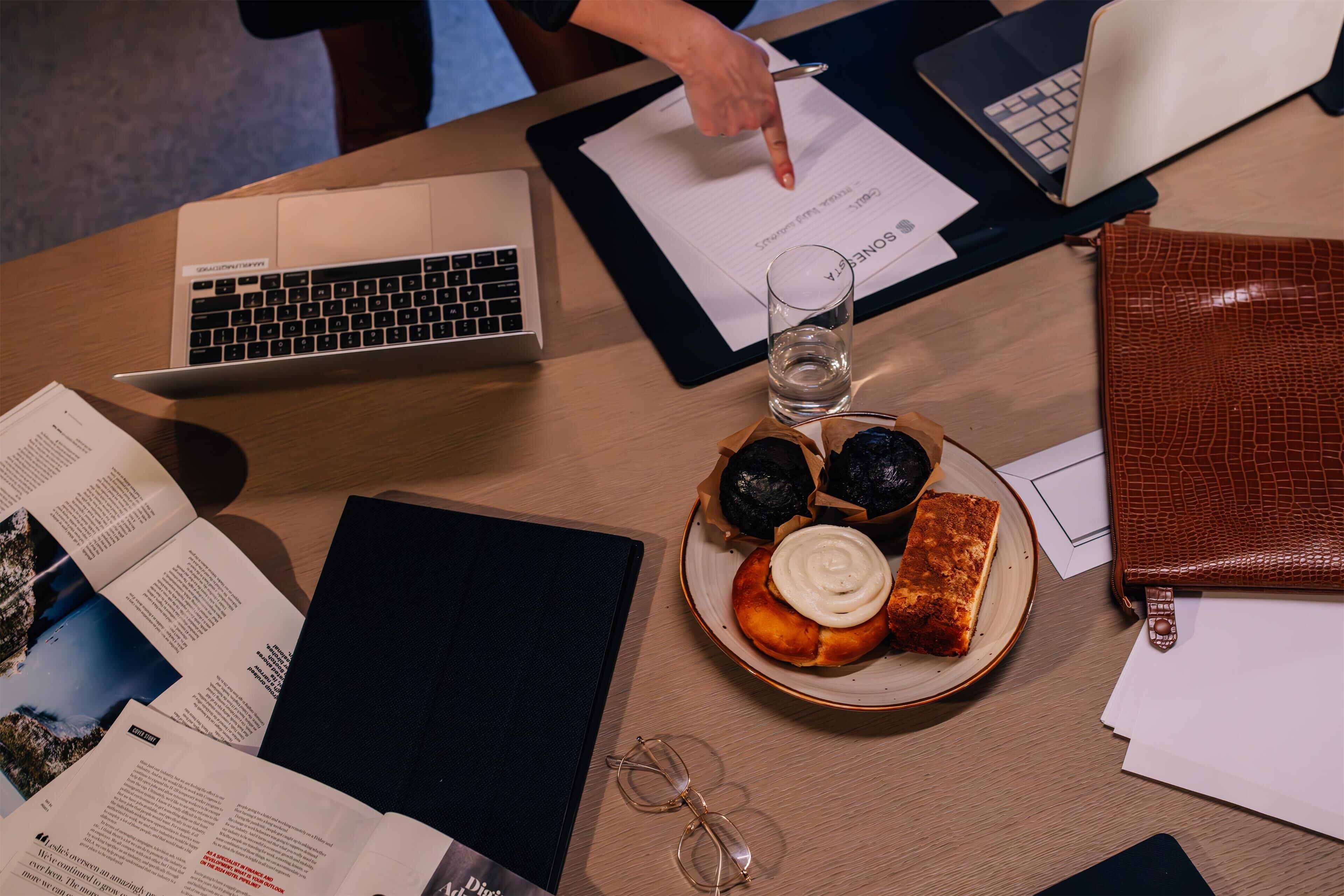 Una mesa con una computadora portátil, documentos, un plato de comida, un vaso de agua y un par de lentes.