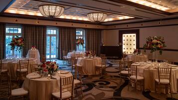 A banquet hall with round tables, chairs, chandeliers, and floral centerpieces.