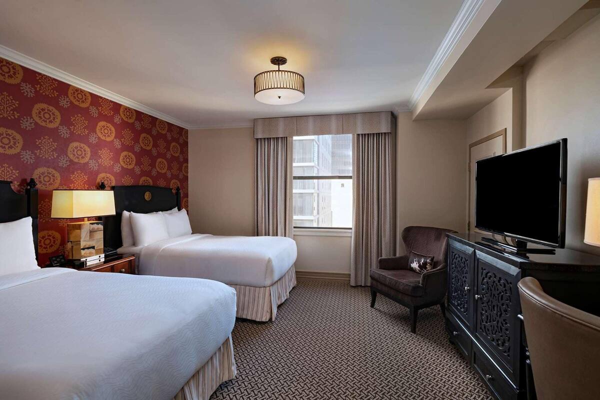Habitación de hotel con dos camas, un televisor, un escritorio con lámpara, una silla y una alfombra estampada.