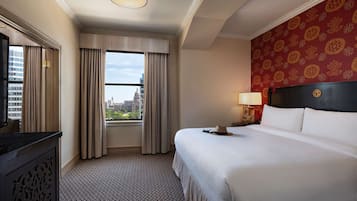 Habitación de hotel con una cama grande, una mesita de noche con lámpara, vista de la ciudad a través de una ventana y un papel tapiz estampado.