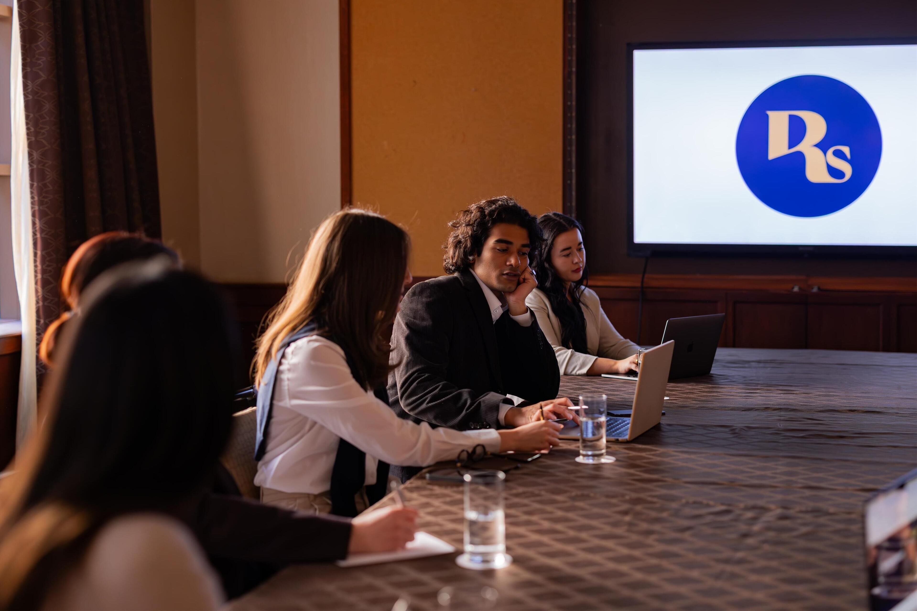 A conference room with a large screen displaying a logo, a long table with laptops and water glasses, and attendees engaged in a meeting.