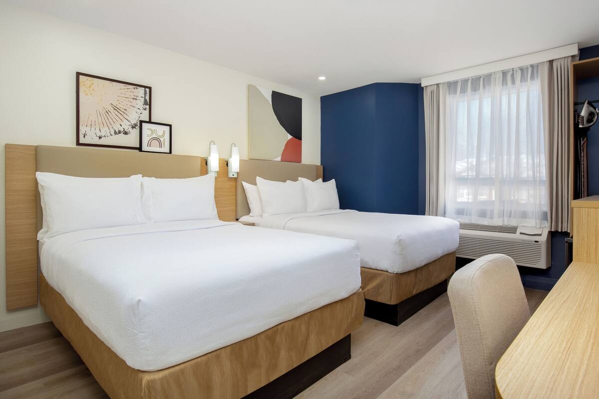 Une chambre d’hôtel avec deux lits, une tête de lit en bois, un bureau et une grande fenêtre avec des rideaux légers.