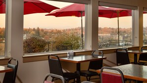 A dining area with tables and chairs, large windows with red umbrellas, and a view of a cityscape with autumn trees.