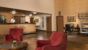 A hotel lobby with a reception desk, red chairs, a small table with a vase, and a wall-mounted sign.