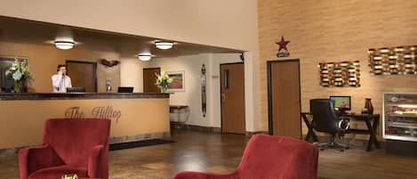 Un vestíbulo de hotel con mostrador de recepción, sillas rojas, una pequeña mesa con un jarrón y un letrero en la pared.
