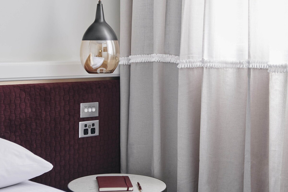 Habitación de hotel con una cama, una mesita de noche con un libro y un bolígrafo, una lámpara y cortinas.