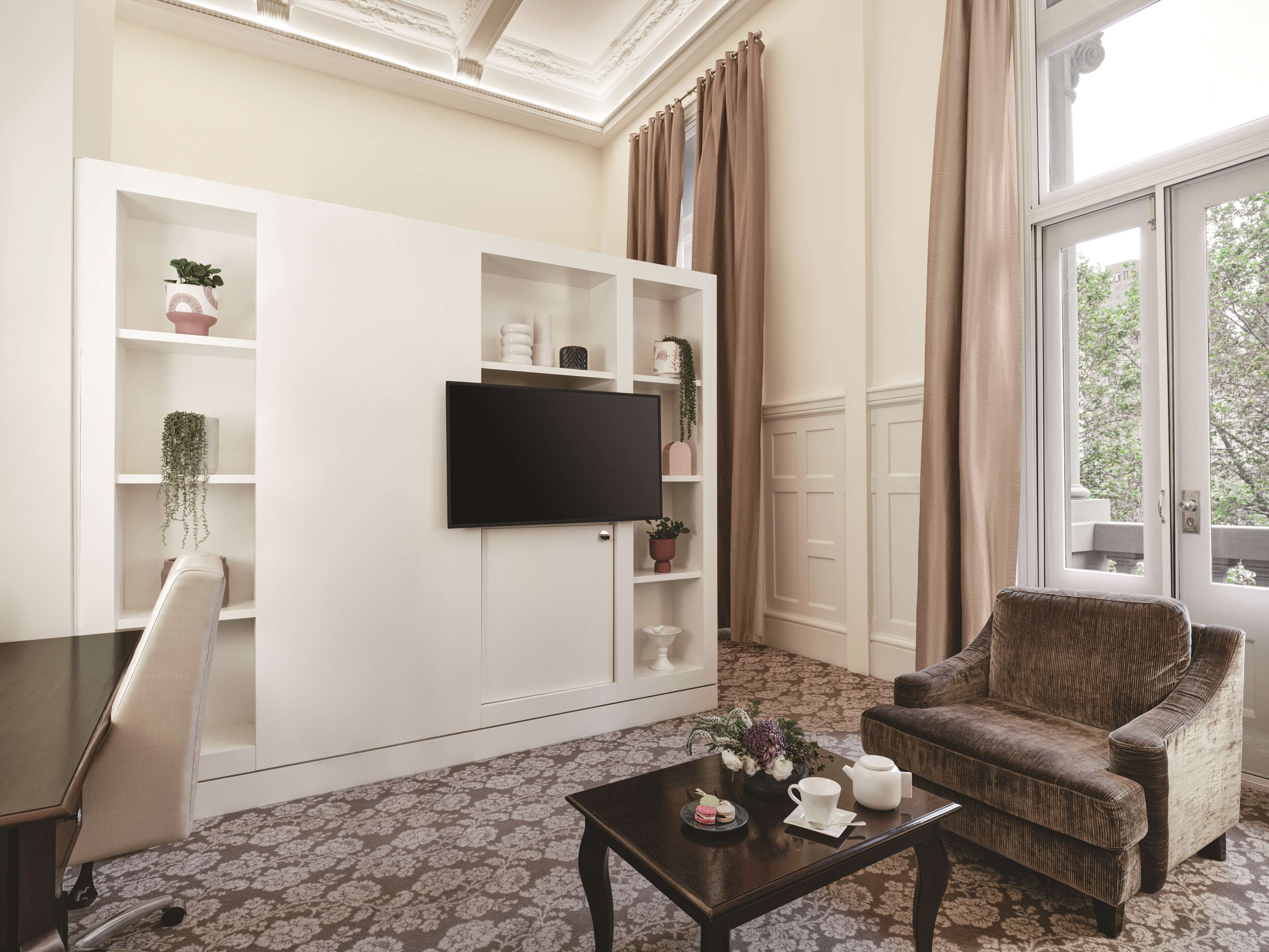 A living room with a flat-screen TV mounted on a white cabinet, a brown armchair, a coffee table with a floral-patterned rug, and a window with curtains.