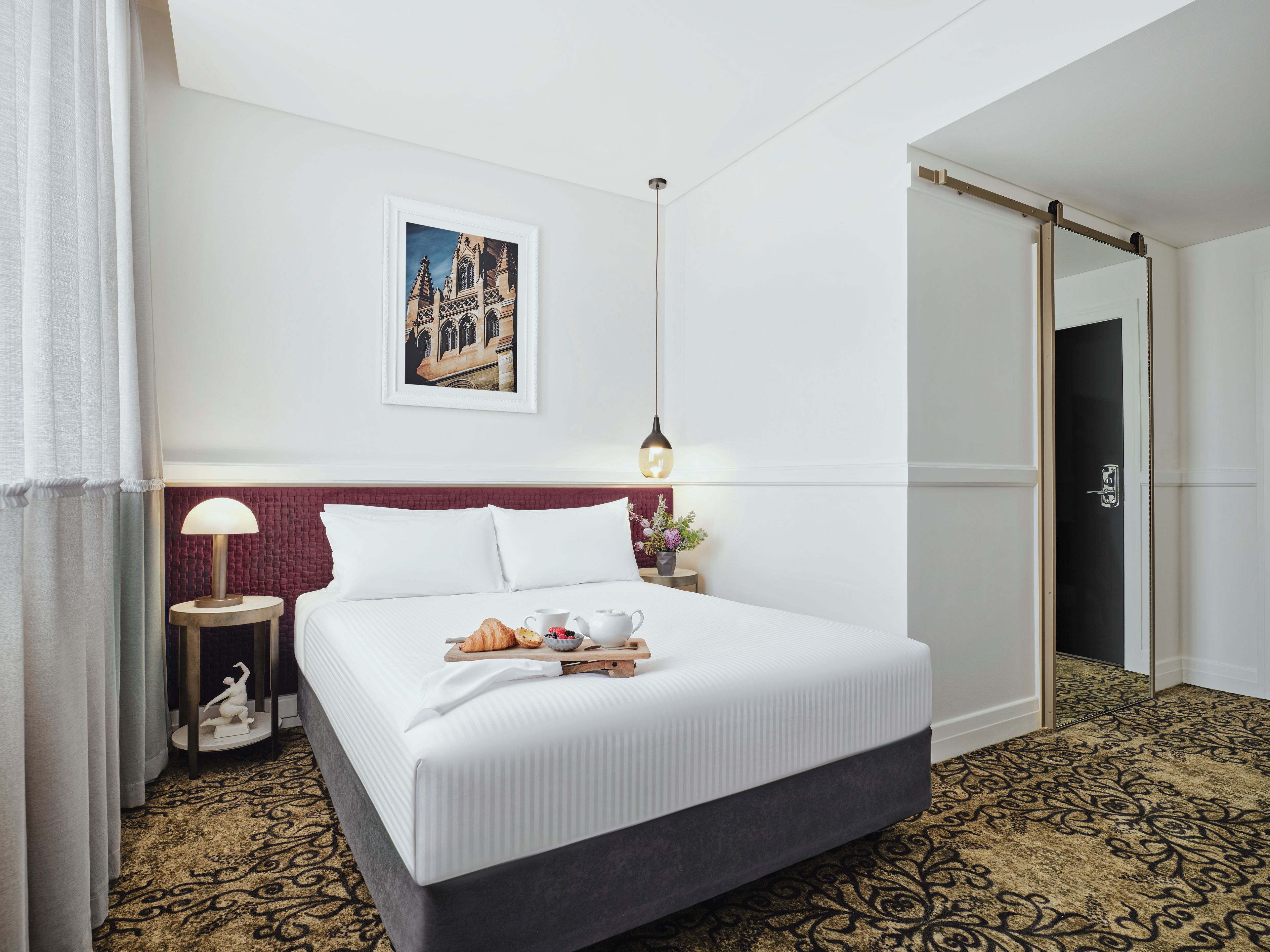 Habitación de hotel con cama, mesita de noche, lámpara, un cuadro en la pared y una puerta.