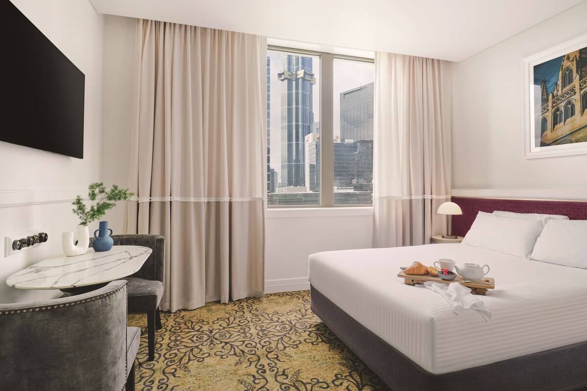A hotel room with a bed, a desk with a vase and plant, a window with curtains, and a painting on the wall.