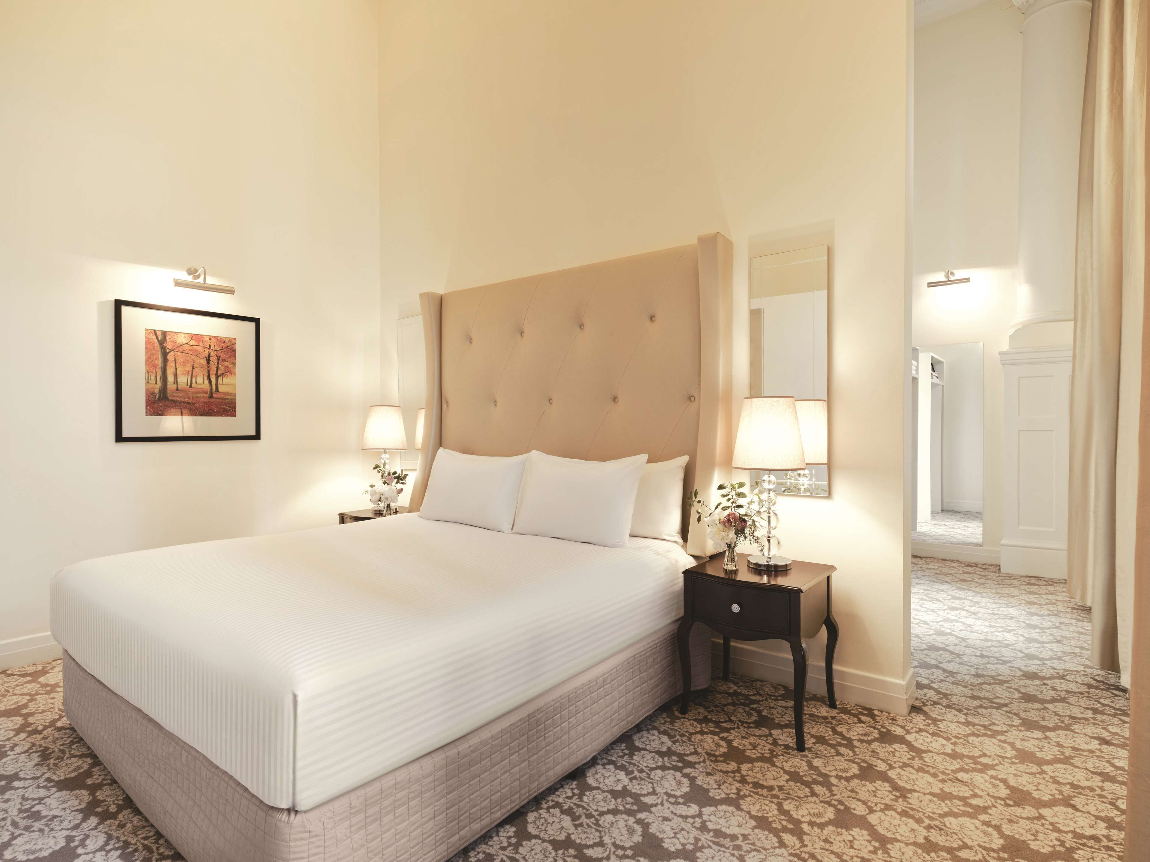 A large bed with a tufted headboard, two bedside tables with lamps, a framed picture on the wall, and a patterned carpet.