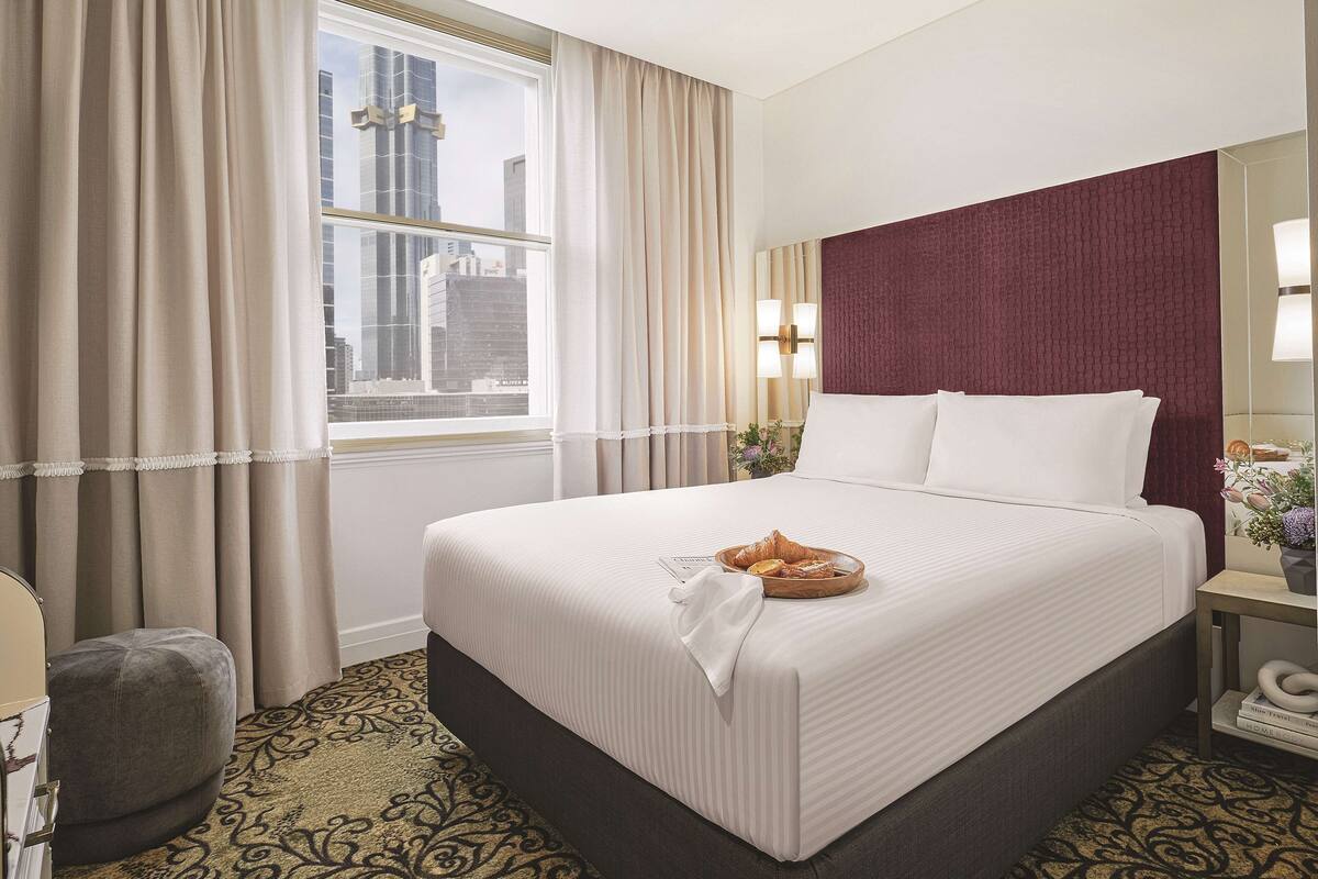 A hotel room with a large bed, a window with curtains, a nightstand with a lamp, and a view of the cityscape.