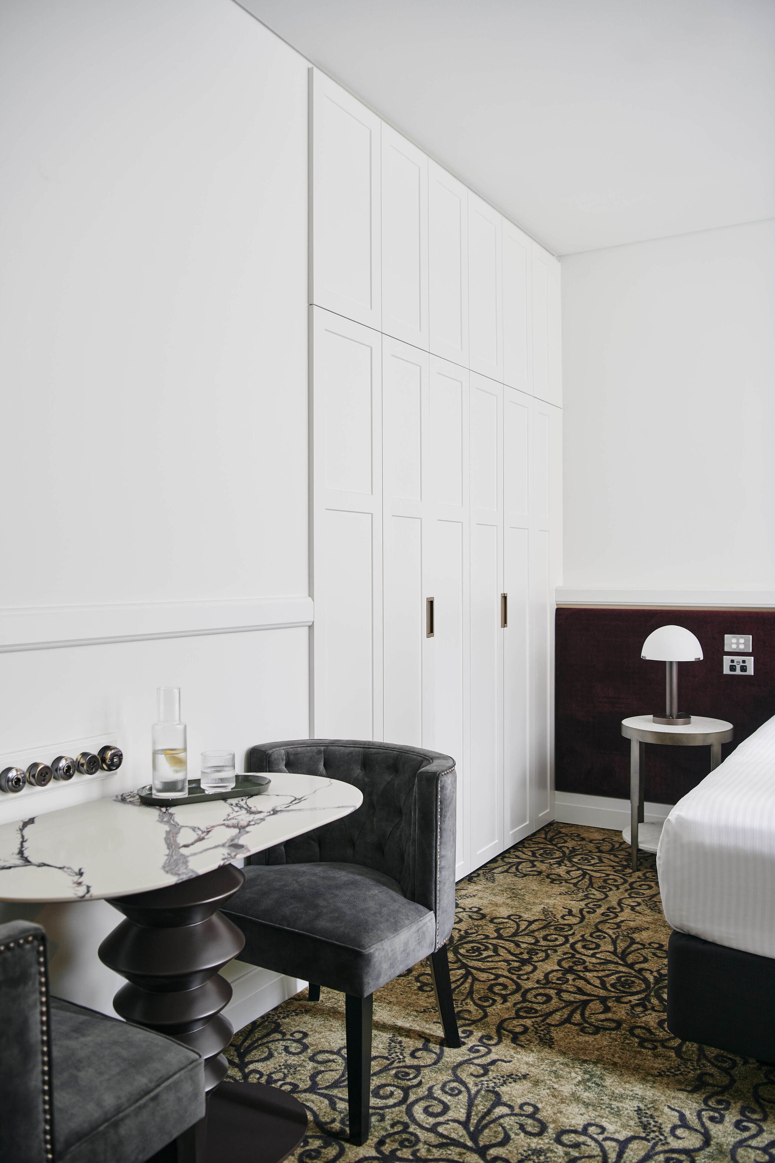 A hotel room with a marble table, a black upholstered chair, a patterned carpet, a bed, and a nightstand with a lamp.