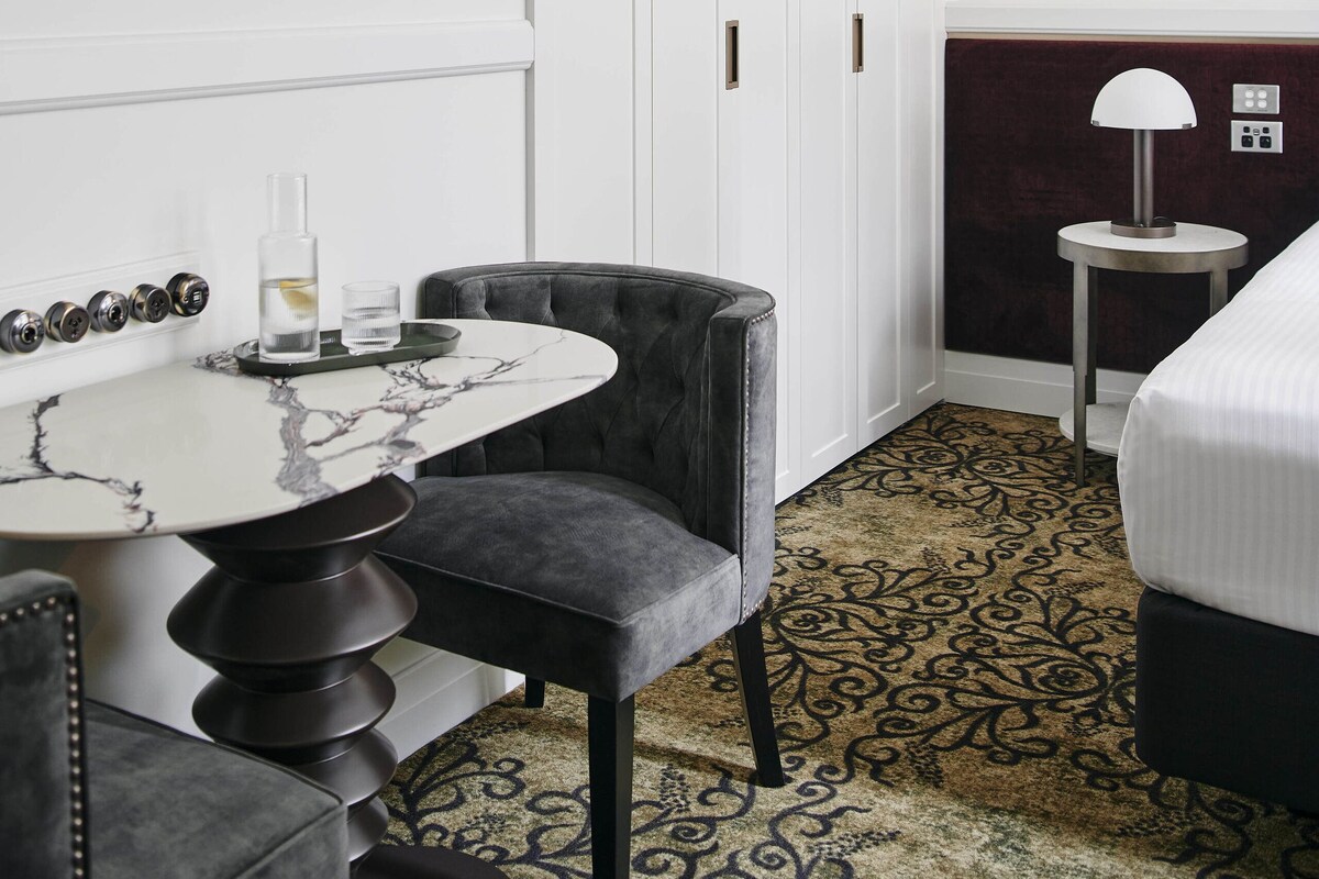 Una habitación de hotel con una mesa de mármol, una butaca tapizada de negro, una alfombra estampada, una cama y una mesita de noche con una lámpara.