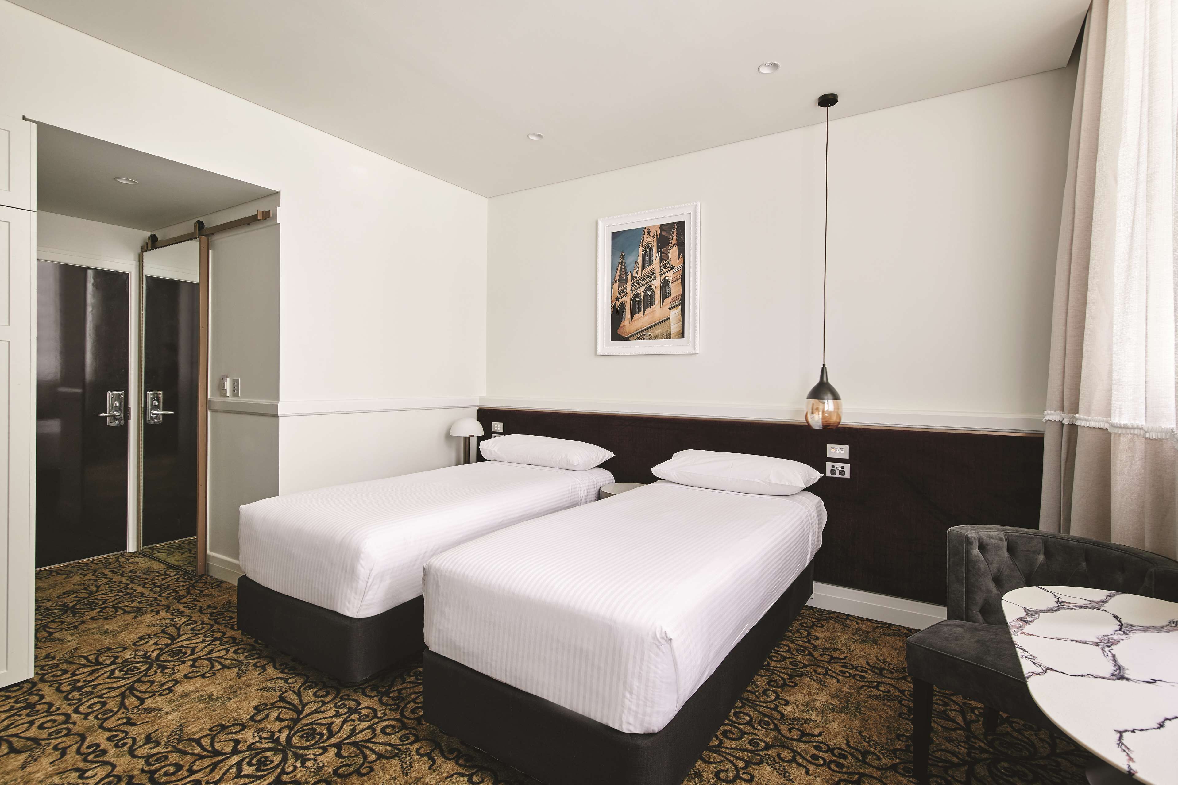 Habitación de hotel con dos camas individuales, un escritorio, una silla, una mesita de noche, una lámpara, un cuadro en la pared y una puerta al baño.
