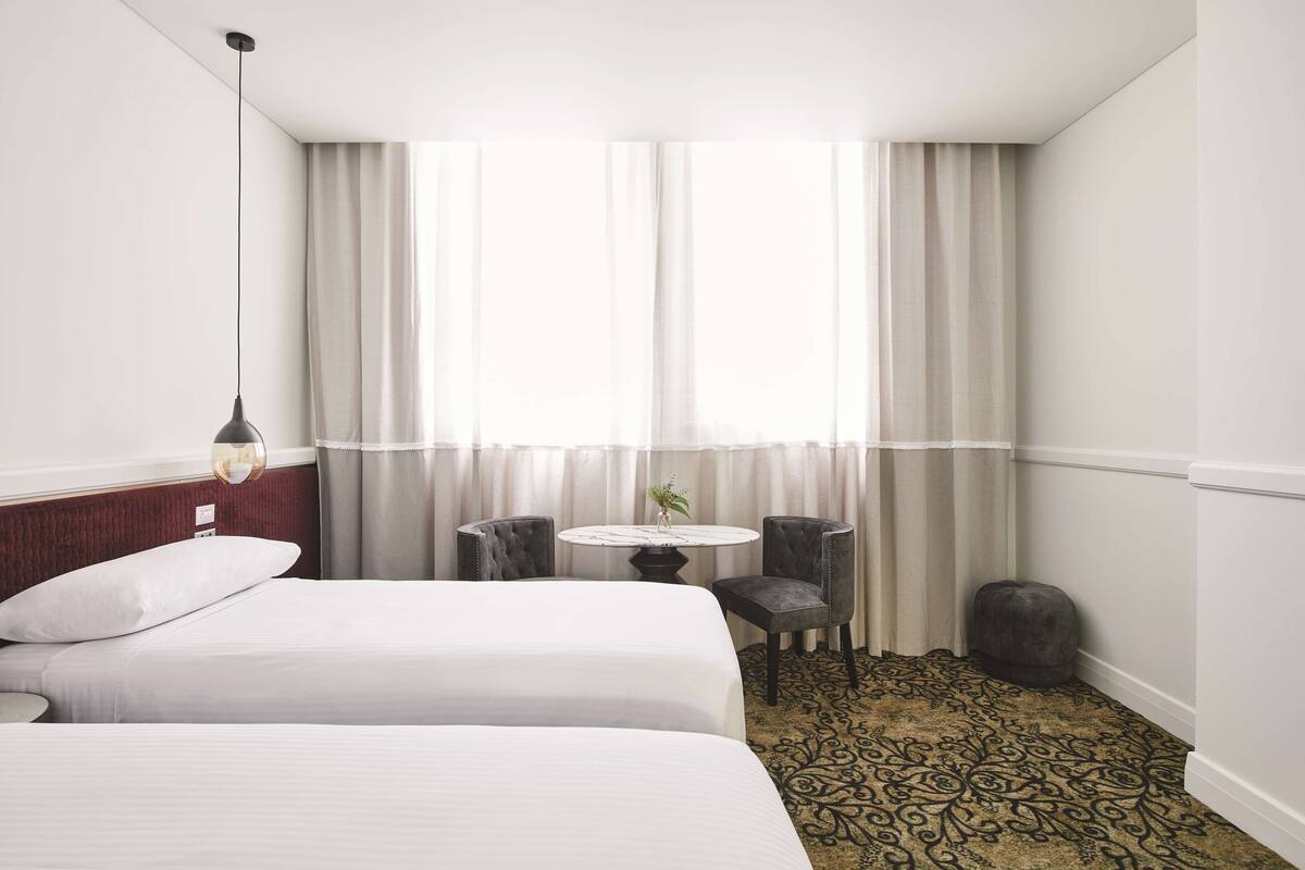 A hotel room with two beds, a small round table with a vase, two chairs, a round ottoman, and a patterned carpet.