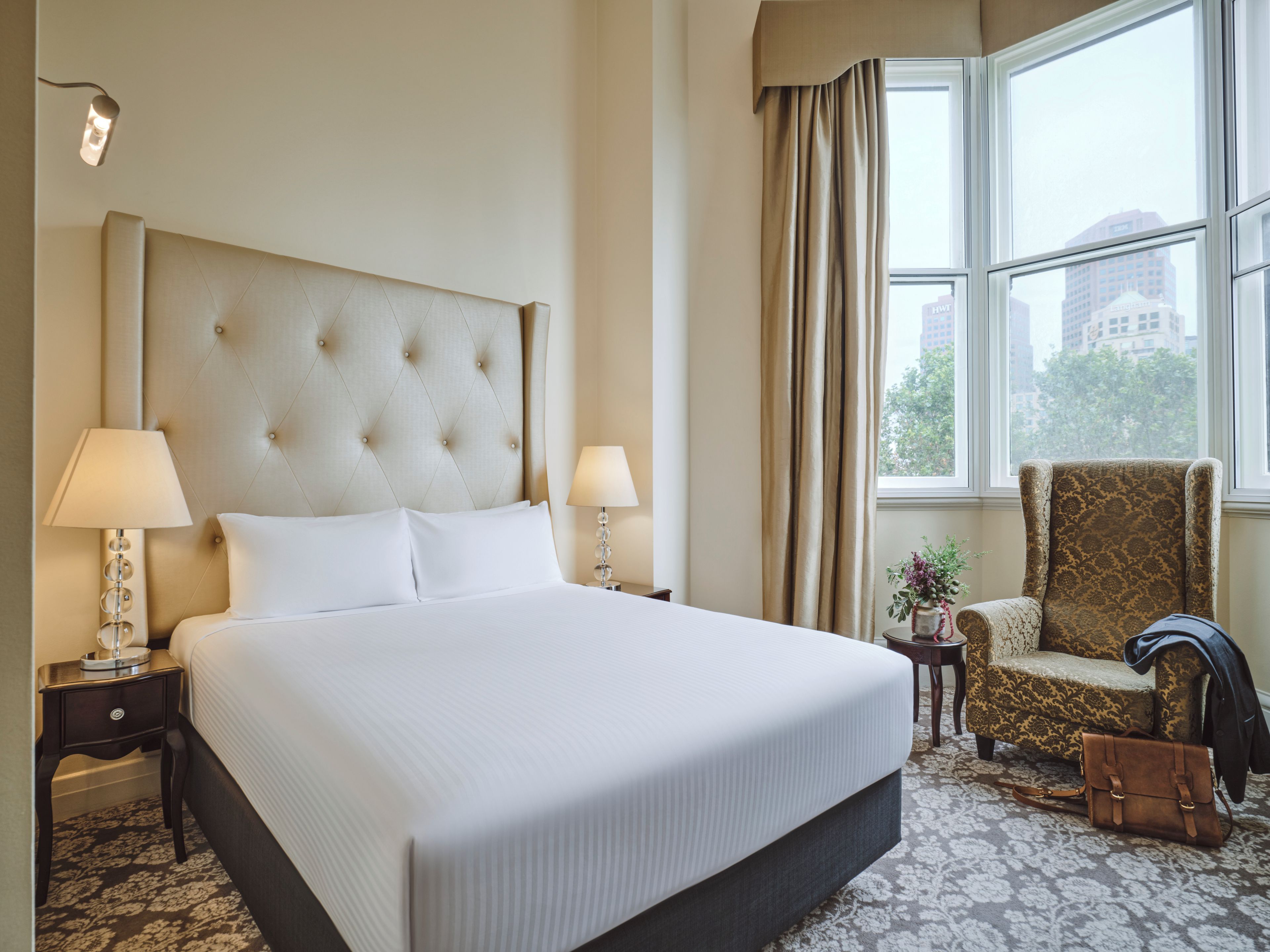 A hotel room with a large bed, two bedside lamps, a chair, a small table, and a window with curtains.