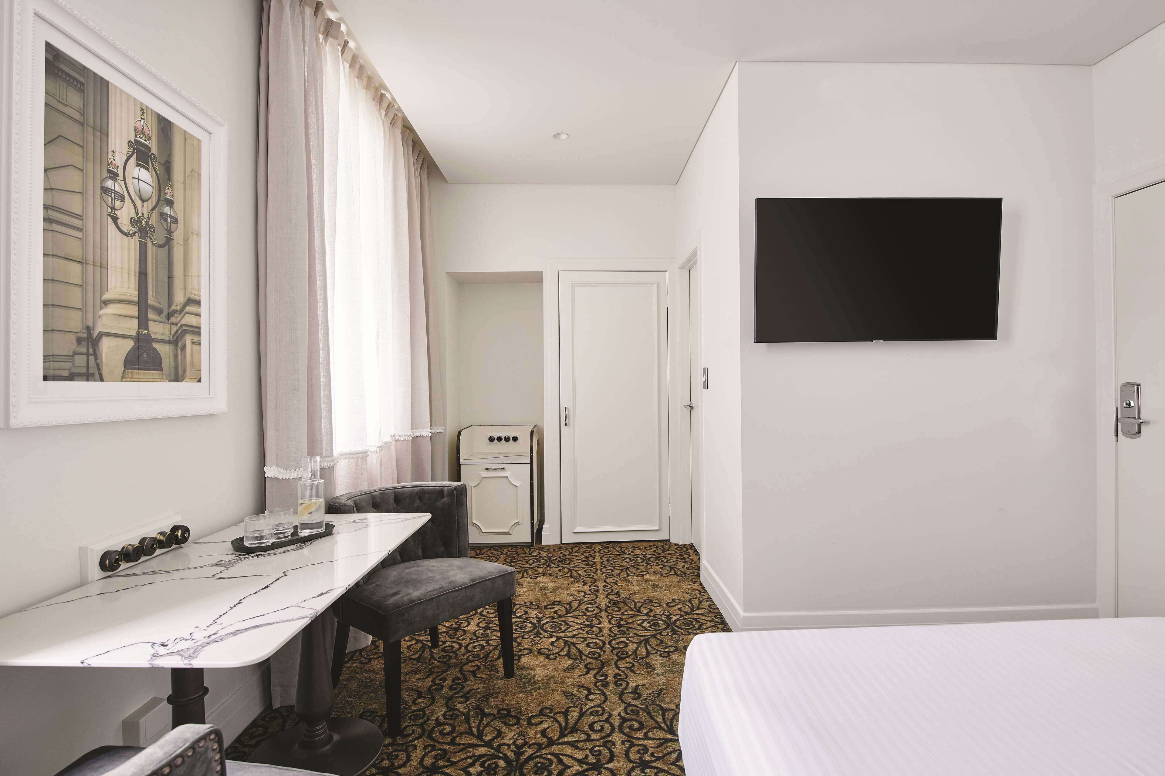 A hotel room with a bed, a desk with a chair, a flat-screen TV mounted on the wall, a wardrobe, and a patterned carpet.