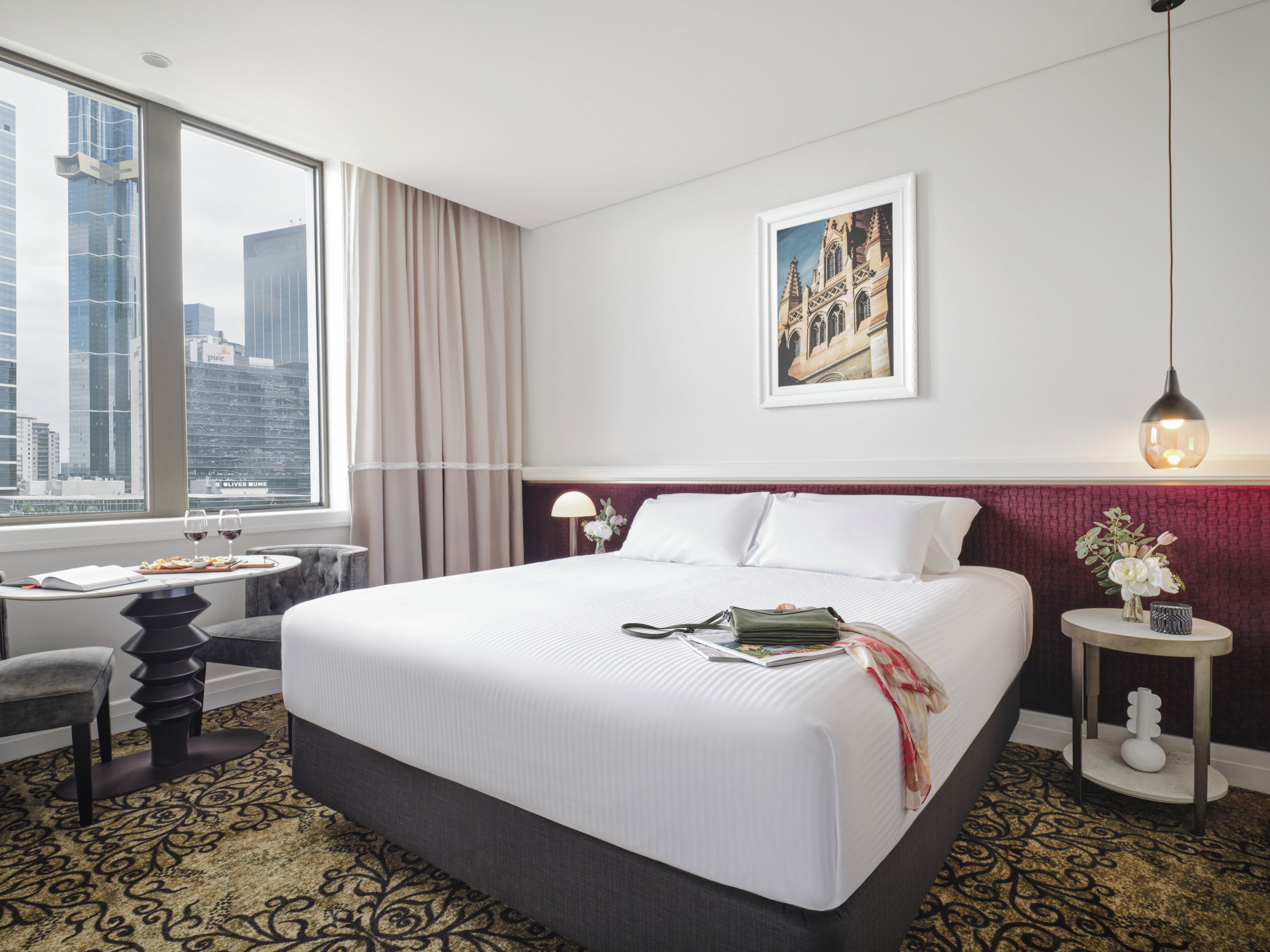 Habitación de hotel con una cama grande, un escritorio con silla, una ventana con cortinas, un cuadro enmarcado en la pared y una mesita con un jarrón de flores.